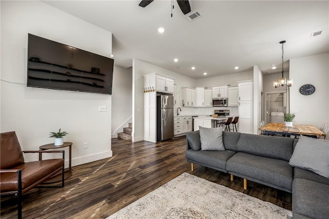 living area featuring ceiling fan with notable chandelier, stairway, dark wood-style floors, and visible vents