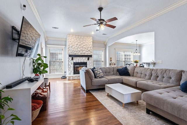 living room with visible vents, a ceiling fan, wood finished floors, a stone fireplace, and crown molding