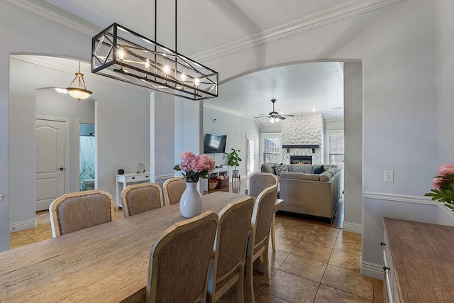 dining space featuring a ceiling fan, arched walkways, ornamental molding, a stone fireplace, and tile patterned flooring