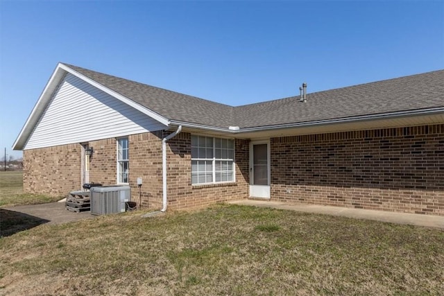 doorway to property featuring brick siding, central AC unit, a shingled roof, and a yard