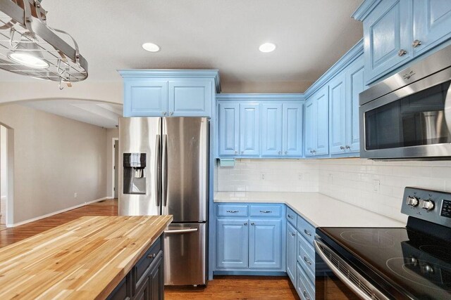 kitchen featuring blue cabinetry, arched walkways, stainless steel appliances, decorative backsplash, and butcher block counters