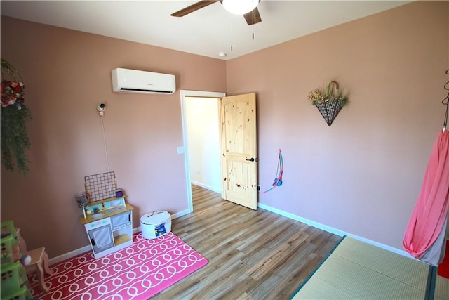 recreation room with wood finished floors, a ceiling fan, baseboards, and a wall mounted AC