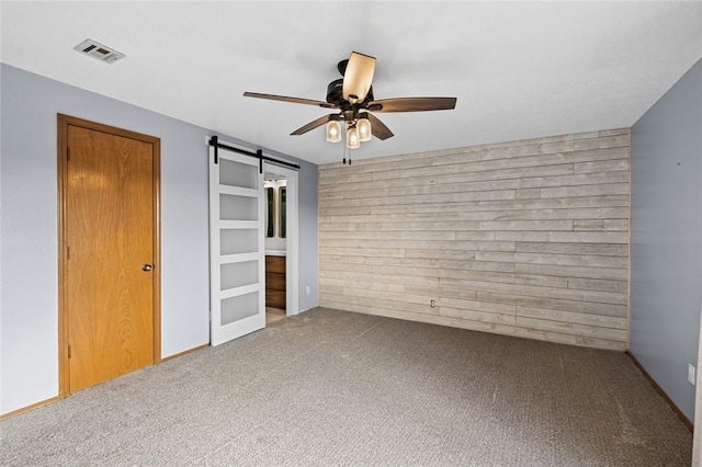 unfurnished room featuring wooden walls, visible vents, carpet floors, ceiling fan, and a barn door