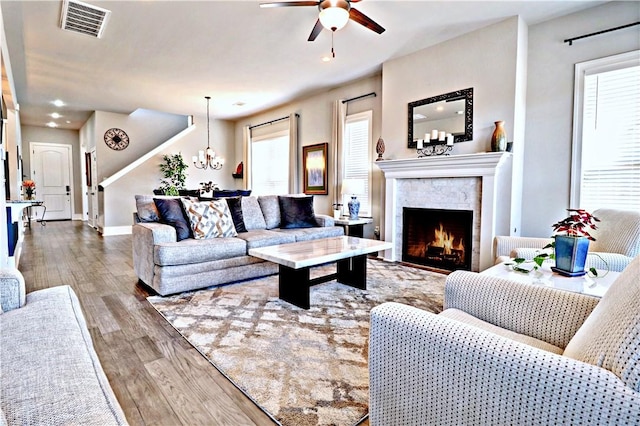 living room with wood finished floors, a wealth of natural light, ceiling fan with notable chandelier, and a warm lit fireplace