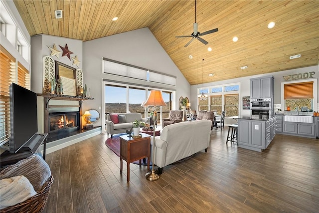 living area with dark wood-type flooring, high vaulted ceiling, a ceiling fan, a glass covered fireplace, and wooden ceiling