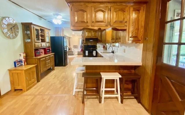 kitchen with brown cabinetry, black refrigerator with ice dispenser, stove, a ceiling fan, and a sink