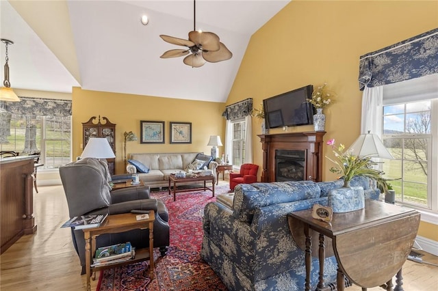 living area featuring high vaulted ceiling, a ceiling fan, wood finished floors, a glass covered fireplace, and baseboards