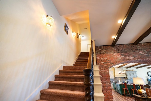 staircase with wood finished floors, arched walkways, beamed ceiling, and brick wall