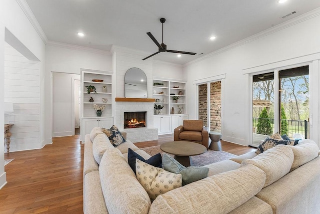 living area with a ceiling fan, crown molding, a brick fireplace, visible vents, and wood finished floors