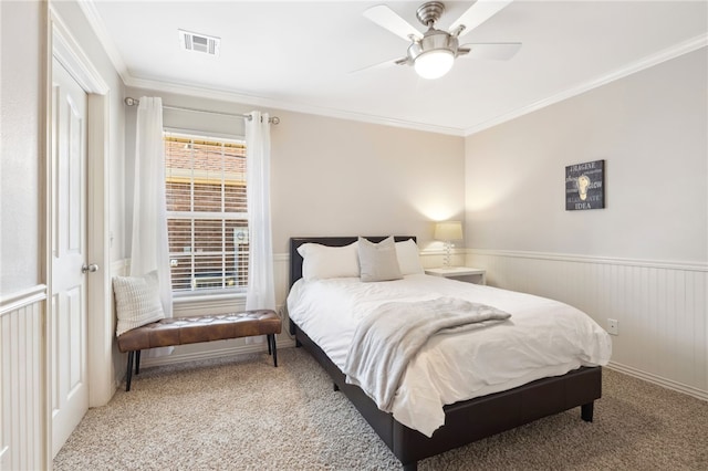 carpeted bedroom with a wainscoted wall, visible vents, ceiling fan, and crown molding