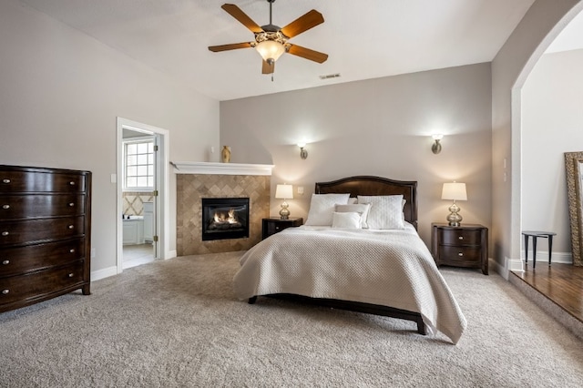 carpeted bedroom featuring a tiled fireplace, baseboards, arched walkways, connected bathroom, and a ceiling fan