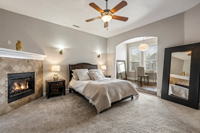 carpeted bedroom featuring a tile fireplace, arched walkways, and a ceiling fan