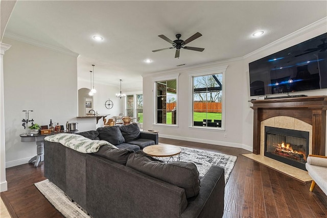 living area with crown molding, a tile fireplace, dark wood-style flooring, and ceiling fan with notable chandelier