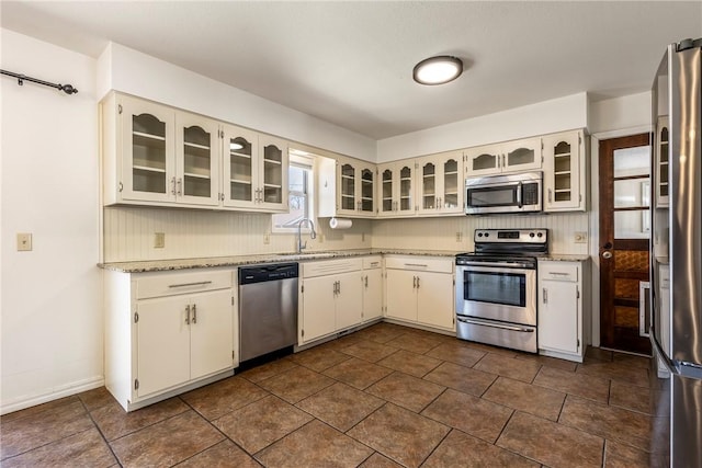 kitchen featuring glass insert cabinets, light stone countertops, a sink, stainless steel appliances, and backsplash