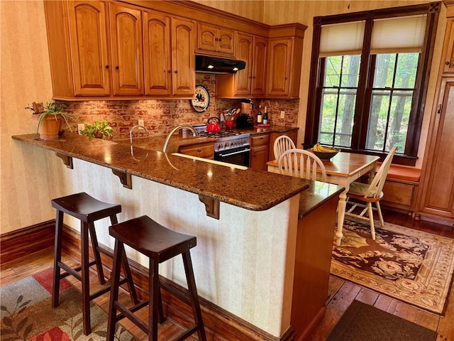 kitchen with brown cabinets, dark stone counters, wallpapered walls, dark wood-type flooring, and under cabinet range hood