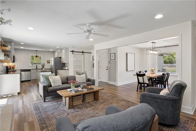 living room with a barn door, wood finished floors, ceiling fan with notable chandelier, baseboards, and recessed lighting