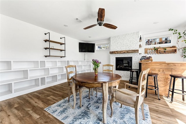 dining room with wood finished floors, visible vents, a ceiling fan, and a large fireplace