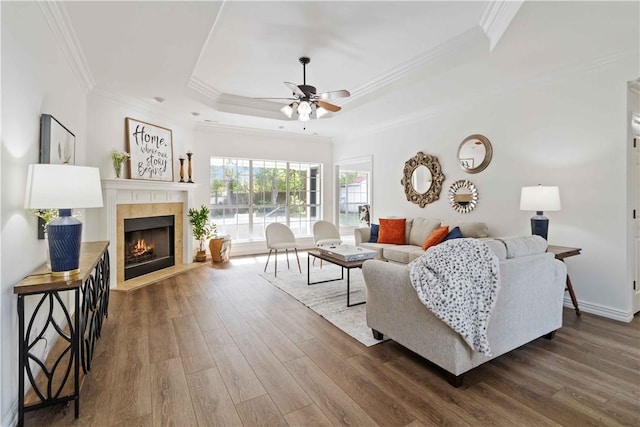 living room with a tray ceiling, a warm lit fireplace, wood finished floors, ornamental molding, and ceiling fan