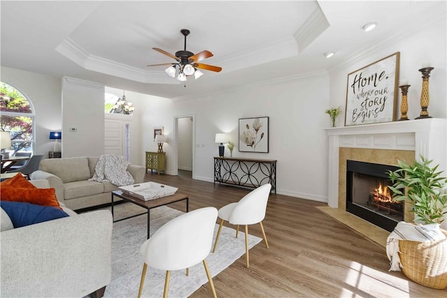 living room featuring a raised ceiling, wood finished floors, ornamental molding, a tiled fireplace, and ceiling fan with notable chandelier