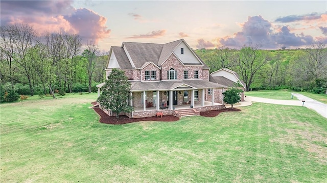 view of front of home with a forest view, a front lawn, a shingled roof, covered porch, and brick siding
