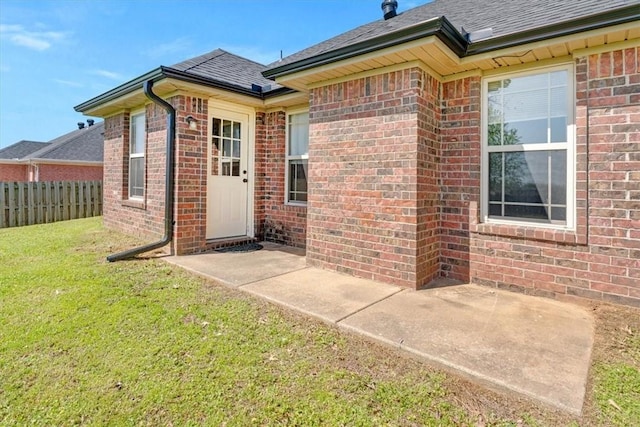 doorway to property with brick siding, a lawn, a shingled roof, and fence