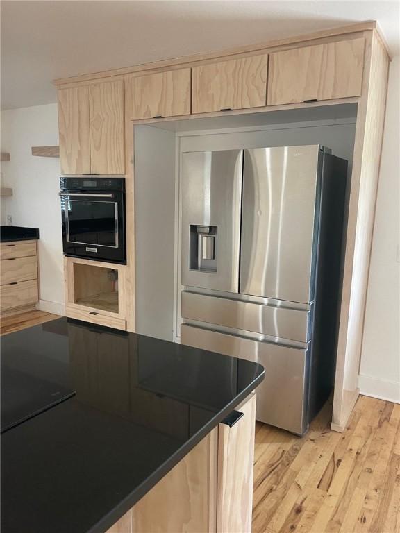 kitchen with stainless steel refrigerator with ice dispenser, dark countertops, light brown cabinetry, and oven