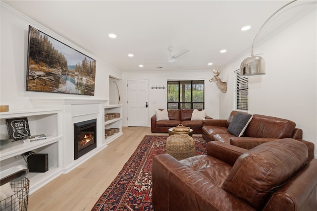 living room with crown molding, ceiling fan, a glass covered fireplace, recessed lighting, and light wood-type flooring