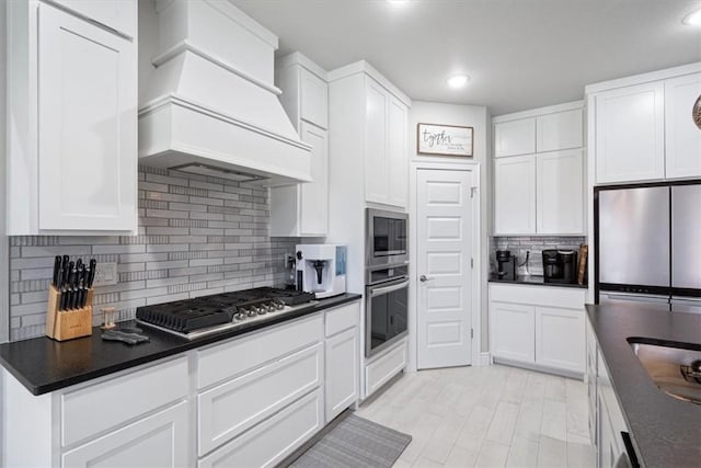 kitchen featuring white cabinets, dark countertops, premium range hood, stainless steel appliances, and backsplash