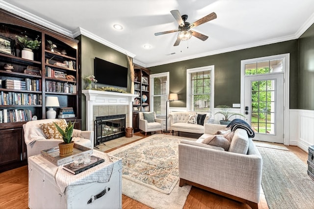 living room featuring wainscoting, a tiled fireplace, a ceiling fan, wood finished floors, and ornamental molding