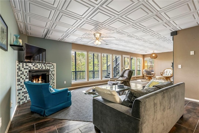 living room with a tiled fireplace, baseboards, ceiling fan, and an ornate ceiling