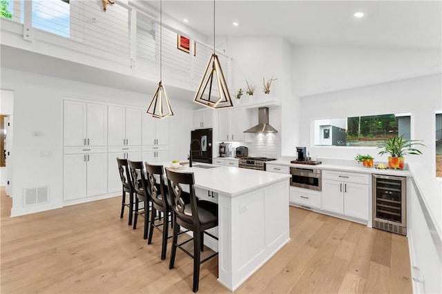 kitchen with visible vents, light wood-type flooring, high vaulted ceiling, wine cooler, and wall chimney exhaust hood