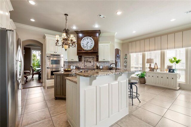 kitchen with a kitchen island with sink, arched walkways, tasteful backsplash, appliances with stainless steel finishes, and ornamental molding