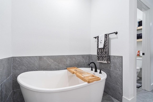 full bath featuring toilet, tile walls, a soaking tub, and a wainscoted wall