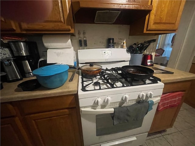 607 Carter St, Clarksville, AR 72830 - kitchen with white gas stove, brown cabinetry, light countertops, light tile patterned floors, and under cabinet range hood
