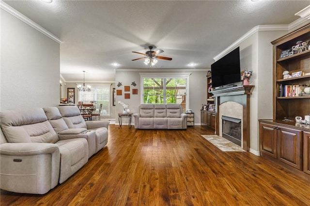 living room with crown molding, dark wood-style flooring, a tiled fireplace, ceiling fan with notable chandelier, and baseboards