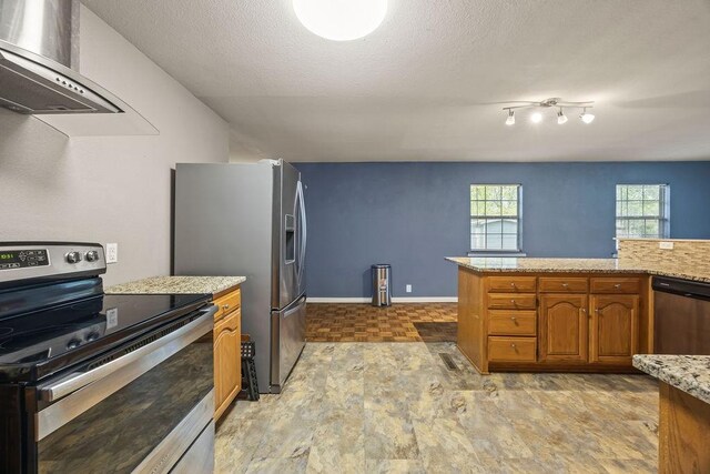 kitchen with a textured ceiling, brown cabinets, light stone counters, stainless steel appliances, and wall chimney exhaust hood