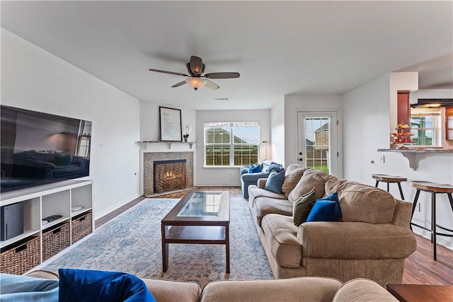 living area with ceiling fan, a tiled fireplace, baseboards, wood finished floors, and visible vents