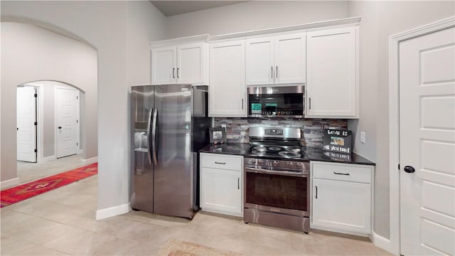 kitchen with dark countertops, appliances with stainless steel finishes, tasteful backsplash, arched walkways, and white cabinetry