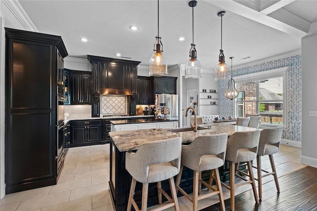 kitchen featuring a sink, a kitchen island with sink, dark cabinetry, tasteful backsplash, and ornamental molding