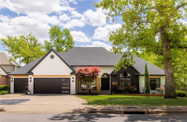 french country style house featuring brick siding, a front lawn, an attached garage, driveway, and roof with shingles