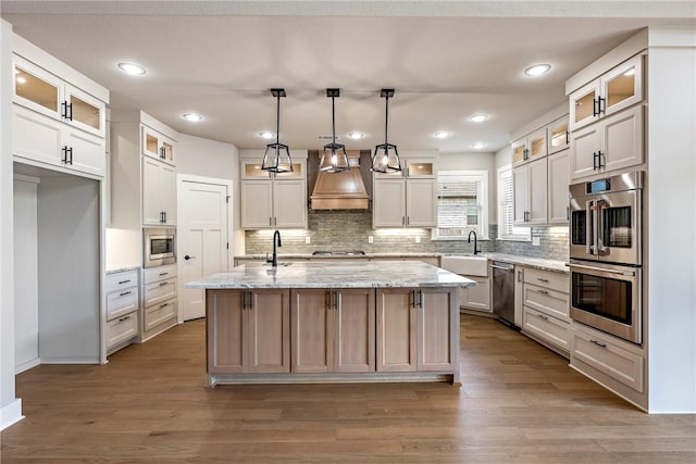 kitchen with a kitchen island with sink, custom exhaust hood, tasteful backsplash, a sink, and appliances with stainless steel finishes