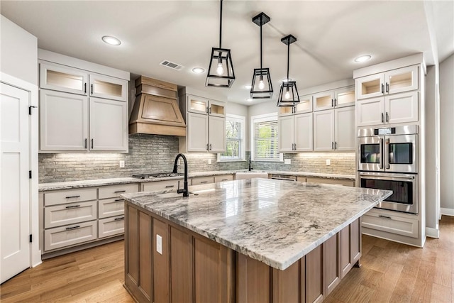 kitchen featuring visible vents, a center island with sink, custom exhaust hood, stainless steel appliances, and light wood-style flooring