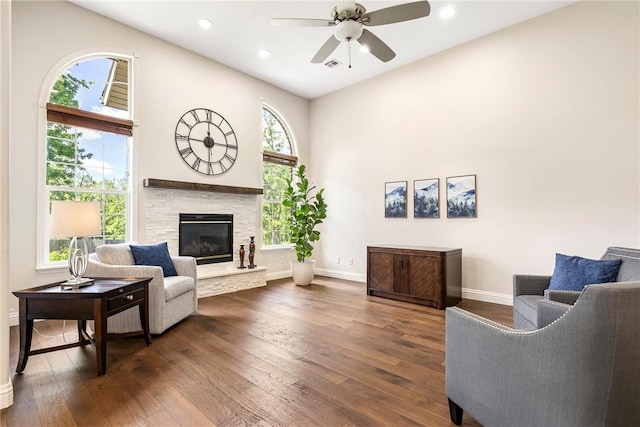 living area featuring a stone fireplace, dark wood-style flooring, baseboards, and ceiling fan