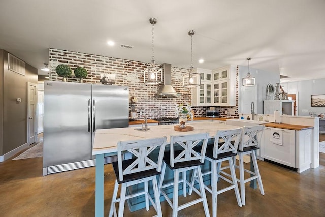 kitchen with freestanding refrigerator, a kitchen island with sink, butcher block countertops, wall chimney exhaust hood, and finished concrete floors
