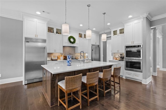 kitchen featuring stainless steel appliances, crown molding, light countertops, glass insert cabinets, and a kitchen island with sink