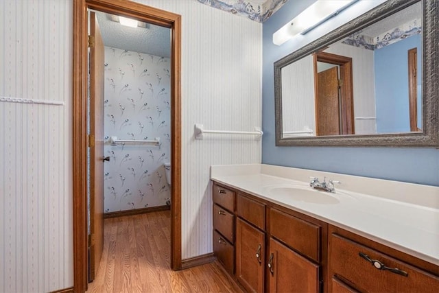 full bath with wood finished floors, vanity, wallpapered walls, toilet, and baseboards