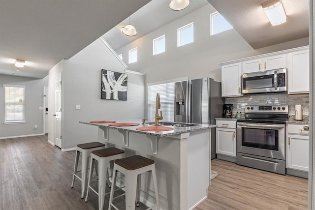 kitchen with stainless steel appliances, decorative backsplash, a kitchen island with sink, light stone counters, and light wood-type flooring