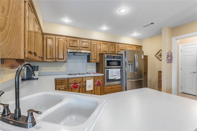 kitchen featuring stainless steel appliances, a sink, light countertops, under cabinet range hood, and brown cabinets