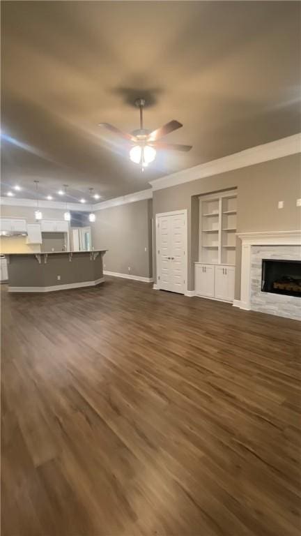 unfurnished living room with ornamental molding, built in shelves, dark wood-style floors, a high end fireplace, and ceiling fan