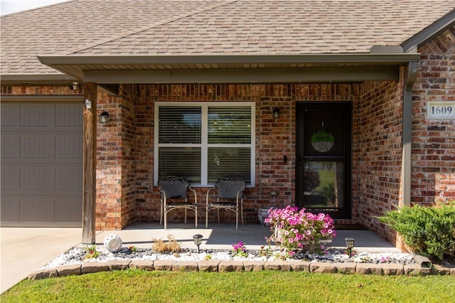 doorway to property featuring roof with shingles, brick siding, an attached garage, and a porch